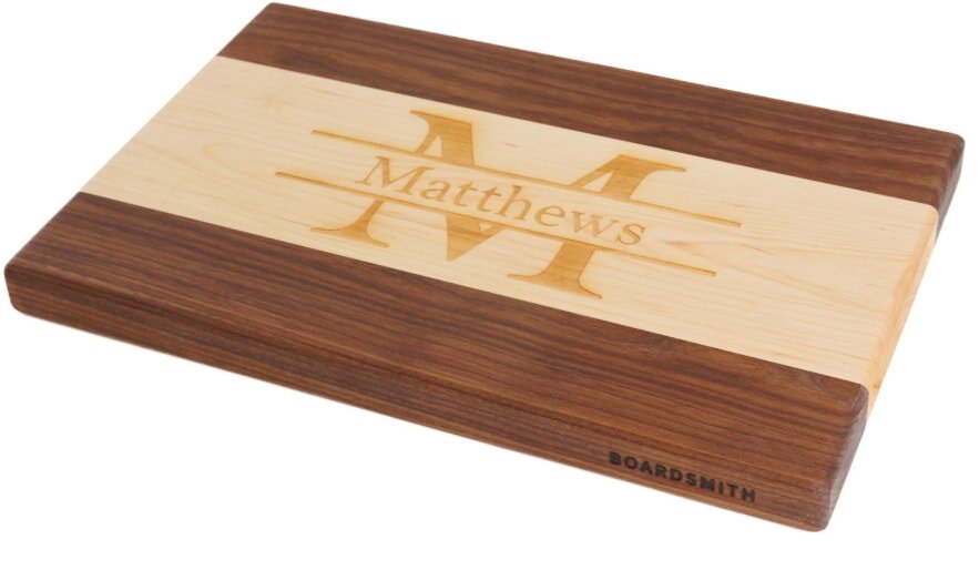 A personalized wooden cutting board with a polished finish features the name "Matthews" and a large "M" engraved in the center. It has a warm, rustic appearance.
