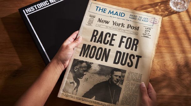 A person holds a newspaper featuring the headline "Rage for Moon Dust," emphasizing a space-related topic.