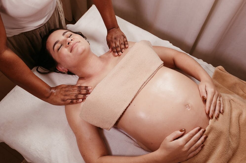 Pregnant woman receiving a relaxing shoulder massage. She lies on her back on a massage table, covered with a towel, eyes closed, conveying calmness.