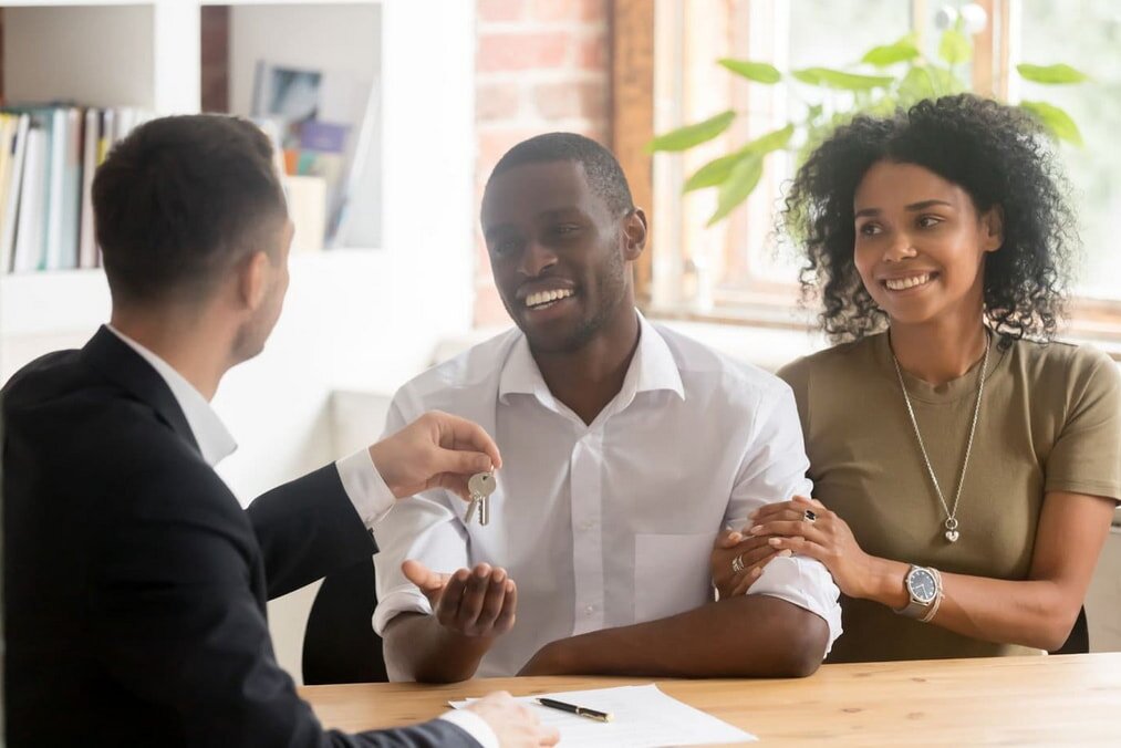 Real estate agent showing a young couple an empty room. The woman in a white blouse holds a tablet and smiles, suggesting a positive, engaging atmosphere.