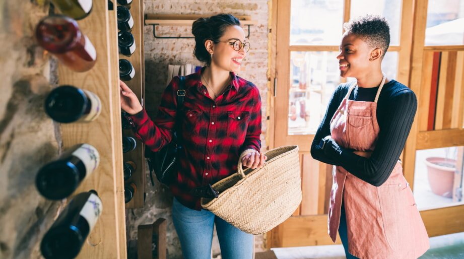 Two women smiling and chatting in a cozy wine shop. One wears a red plaid shirt and glasses, holding a woven bag. Wine bottles line the wooden shelves.