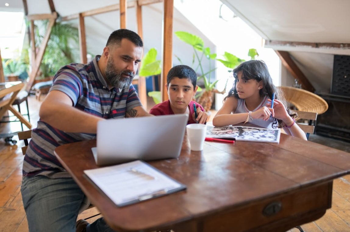 A man and two children are seated at a table, engaging with a laptop in a cozy indoor setting.