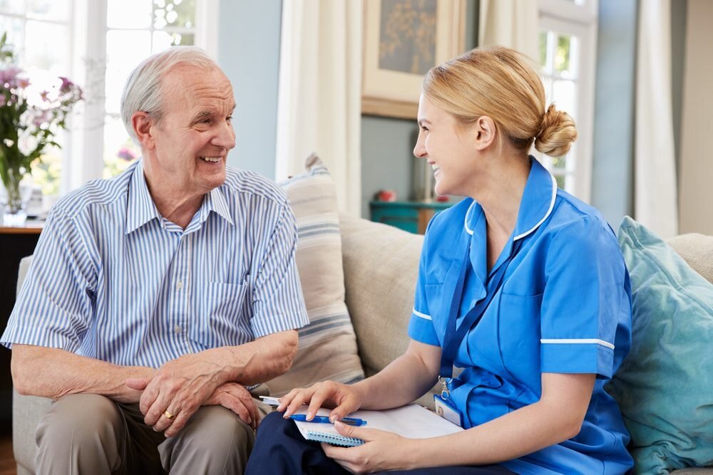 An older man and a nurse are seated on a couch, sharing a moment of conversation in a comfortable environment.