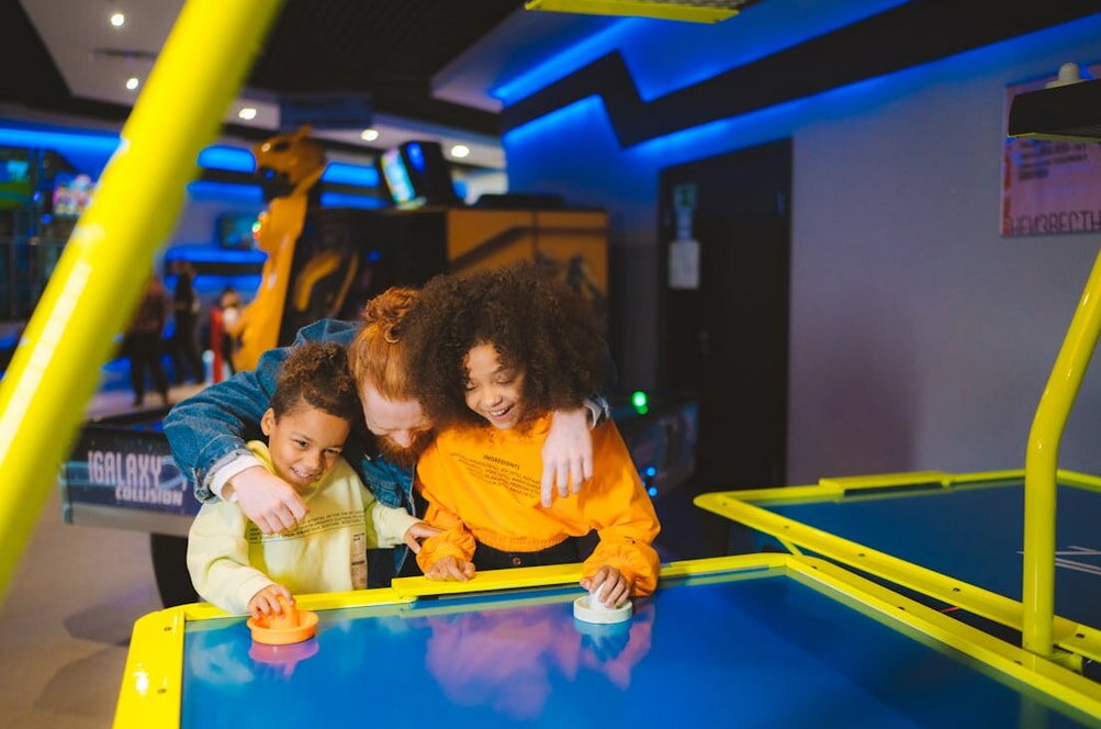 A group of three children happily play air hockey in a vibrant arcade. The neon blue and yellow table and bright lights create an energetic atmosphere.