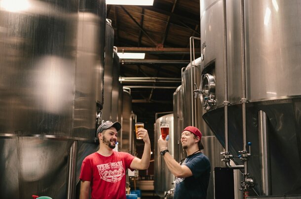 Two men raise their beer glasses in a toast inside a brewery, smiling at each other.