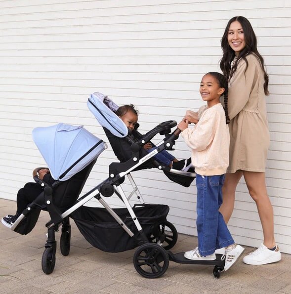 A smiling woman stands with a double stroller on a sidewalk. Two children sit in the stroller, while a third child stands on a board, also smiling.