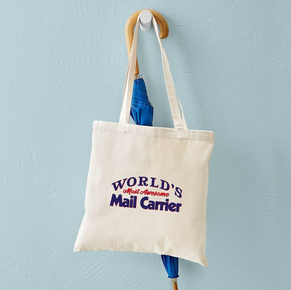 A cream tote bag hangs on a wooden hook against a light blue wall. The bag reads "World's Most Awesome Mail Carrier" in colorful text. A blue umbrella peeks out from beneath. The scene feels cheerful and light-hearted.