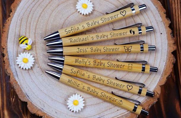 Six engraved wooden pens arranged on a tree slice display board, each with a different name for a baby shower. Decorated with daisies and a bee, conveying a cheerful, celebratory tone.