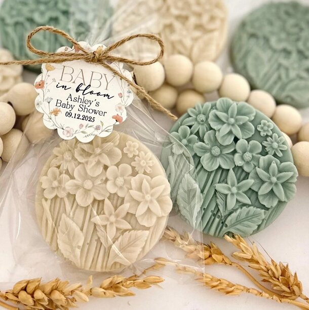 Cream and green floral embossed soaps wrapped in clear packaging with a twine bow. Tag reads "Baby in Bloom, Ashley's Baby Shower, 09.12.2025." Dried wheat accents.