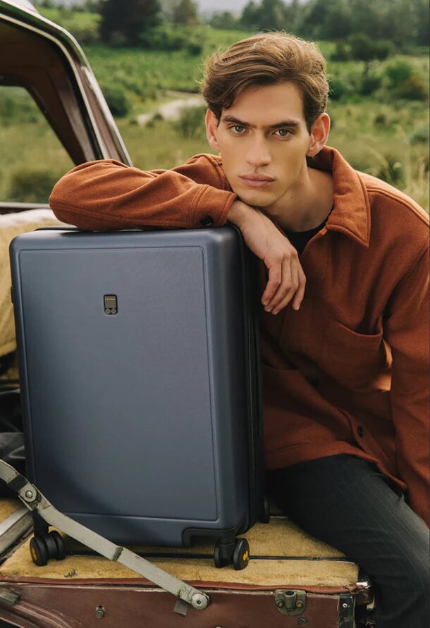 A man sits on a suitcase beside a parked car, looking relaxed and contemplative in an outdoor setting.
