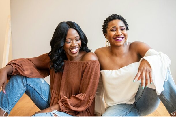Two women sitting on the floor, smiling and laughing together in a joyful moment.