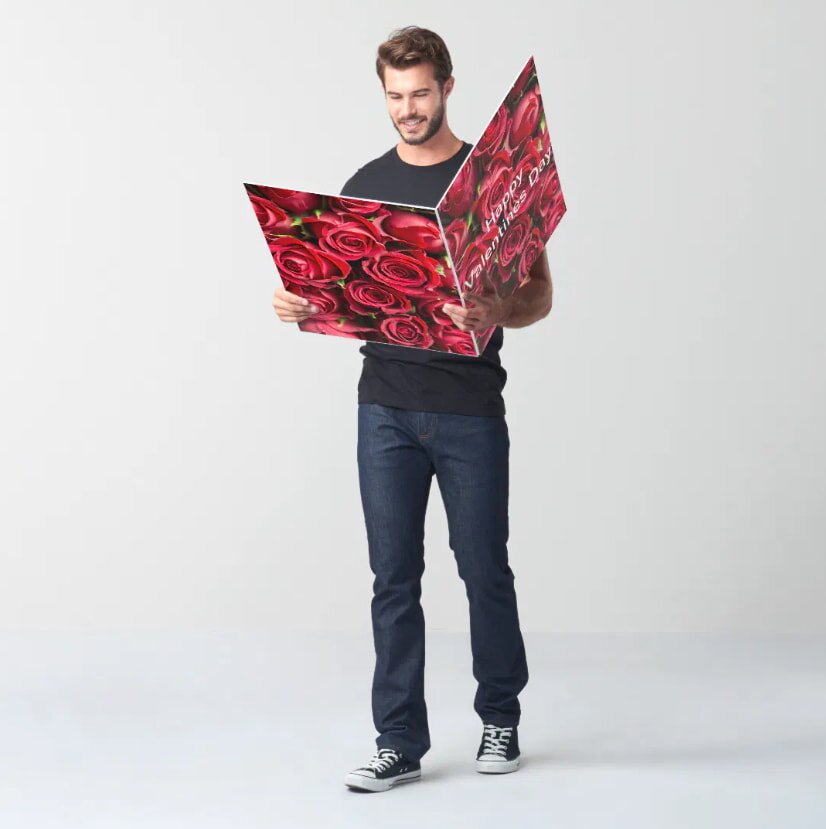 A man in jeans and a t-shirt smiles while holding a large, colorful card featuring red roses and "Happy Valentine's Day" text on a plain background.