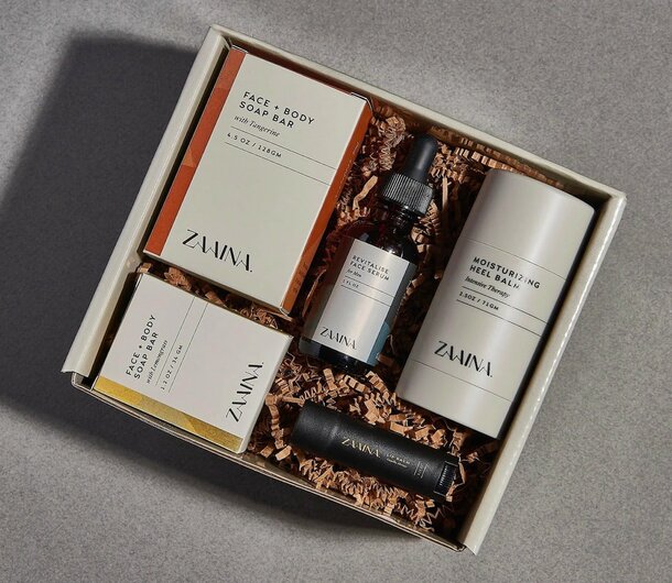 A gift box containing skincare items on shredded paper: two soap bars, a face serum, a moisturizing heel balm, and a lip balm tube, all labeled "Zavina."