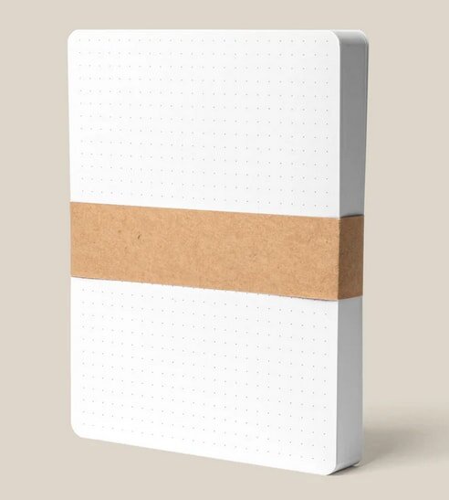 A dotted notebook with a brown paper band stands upright against a beige background, showcasing its minimalist design.