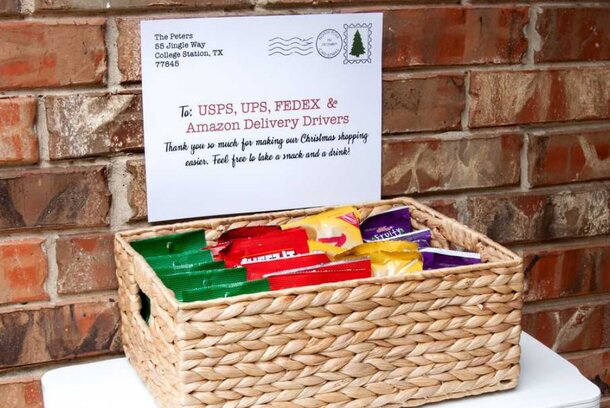 A woven basket filled with snacks is placed against a brick wall. A note reads: "To USPS, UPS, FEDEX & Amazon Delivery Drivers. Thank you! Feel free to take a snack and drink!"