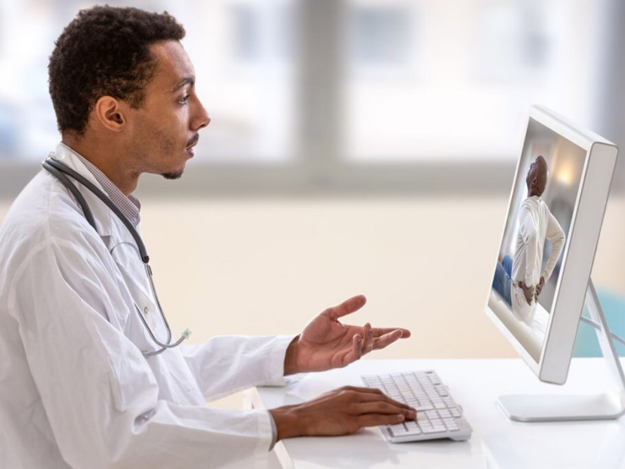 A male doctor sits at a desk, focused on a computer screen, with medical documents and a stethoscope nearby.