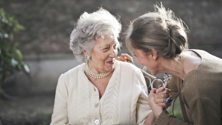 An elderly woman and a younger woman share a joyful moment, smiling and holding hands outdoors. The scene conveys warmth and affection.