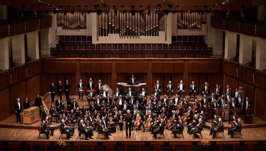 An orchestra on a wooden stage, musicians in formal attire with a conductor at center. A grand pipe organ is visible in the background, creating an elegant and formal atmosphere.