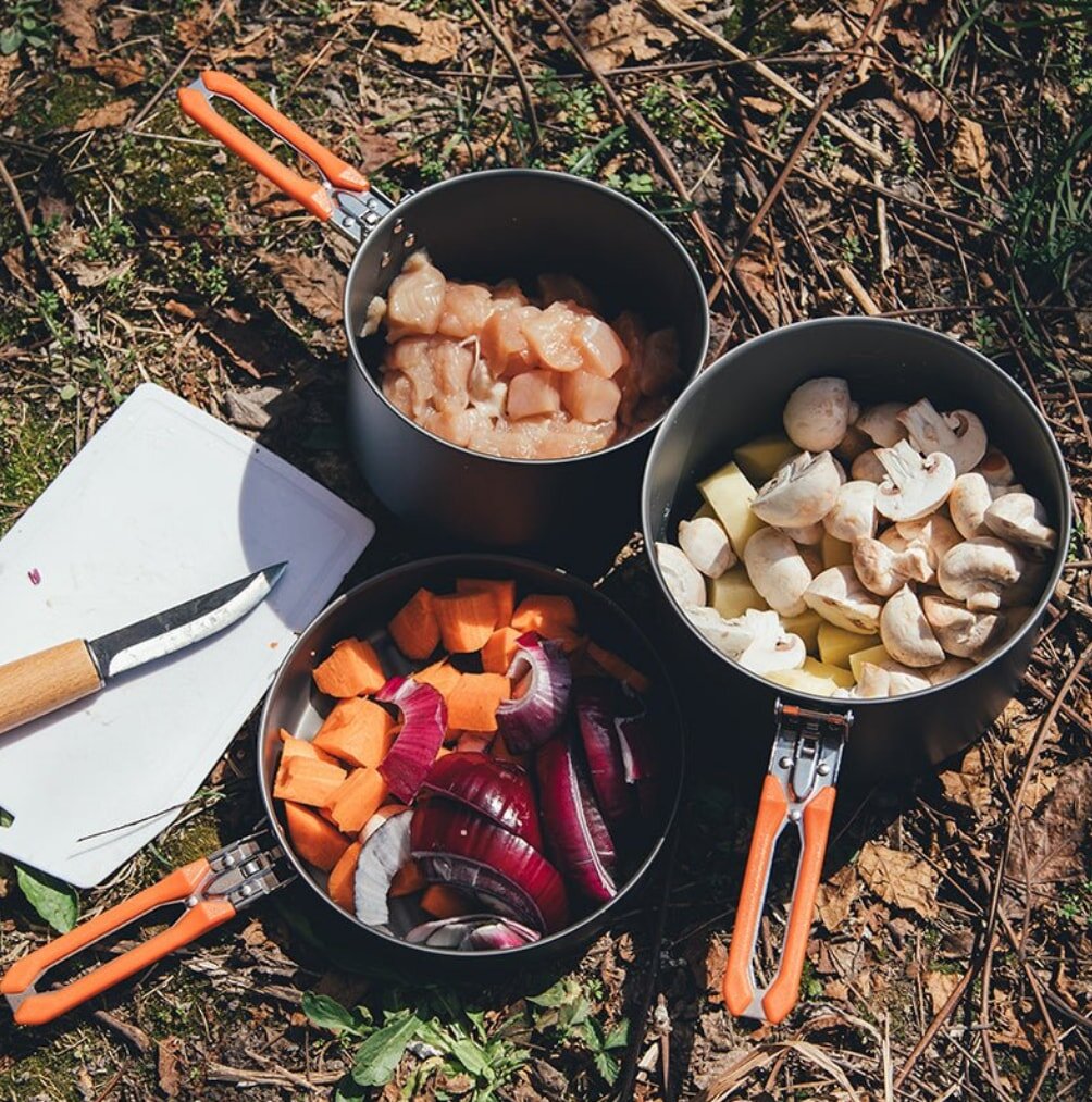 Cooking pots filled with fresh vegetables and mushrooms placed on the ground, ready for meal preparation.