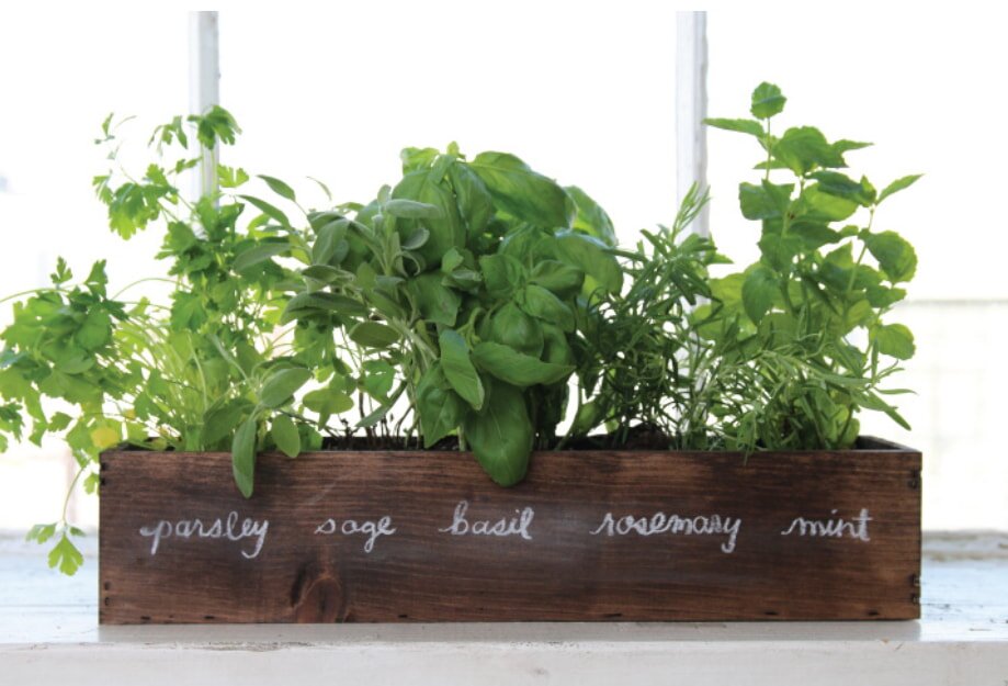 A wooden planter box with "parsley sage basil rosemary mint" written on it holds lush green herbs, set against a bright window, conveying freshness.