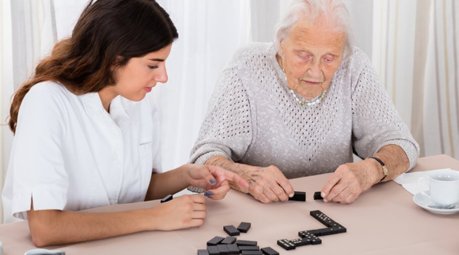 A young woman and an elderly woman sit at a table playing dominoes. Both are focused and engaged, with some dominoes laid out and others scattered.