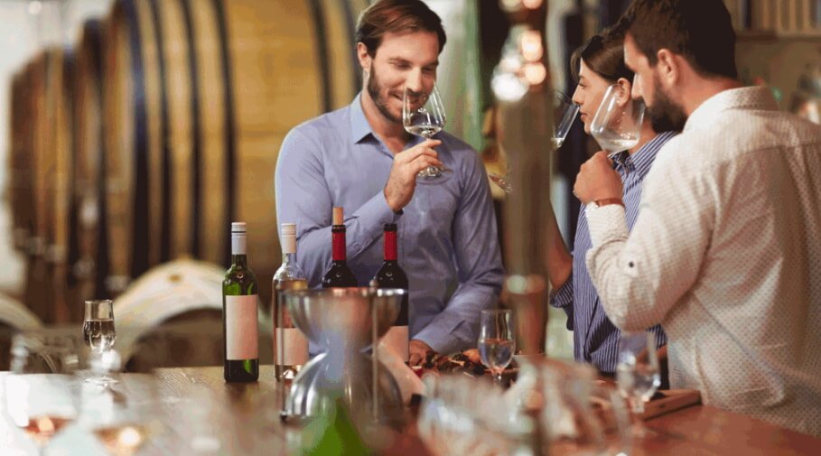 Three people enjoy wine tasting, holding glasses and smiling, in a winery with wooden barrels. Bottles are on the table, creating a warm, convivial atmosphere.