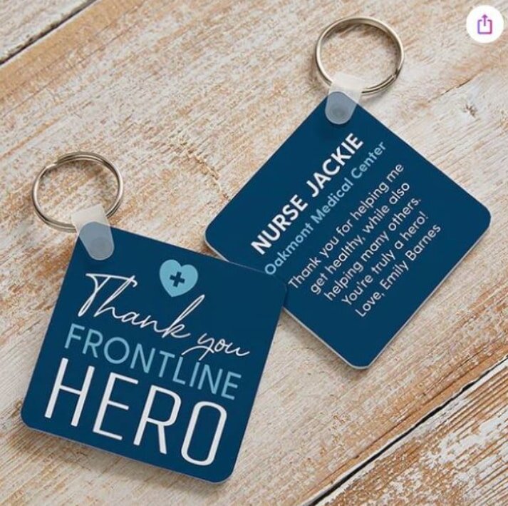 Two blue square keychains on a wooden surface. One reads "Thank you FRONTLINE HERO" with a heart. The other thanks Nurse Jackie from Oakmont Medical Center.