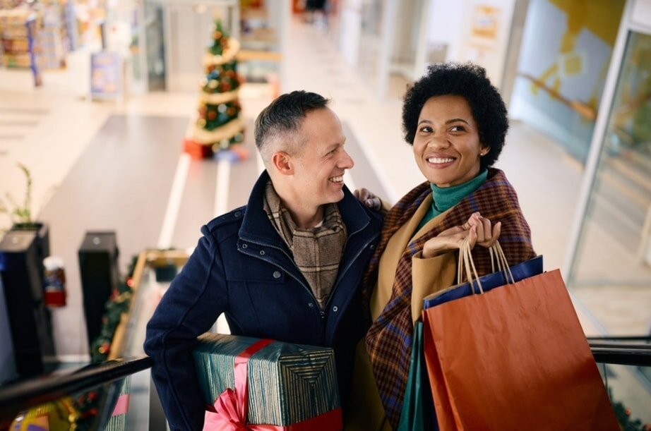 A smiling couple on an escalator hold shopping bags and gift boxes, set in a festive mall with a decorated Christmas tree in the background.