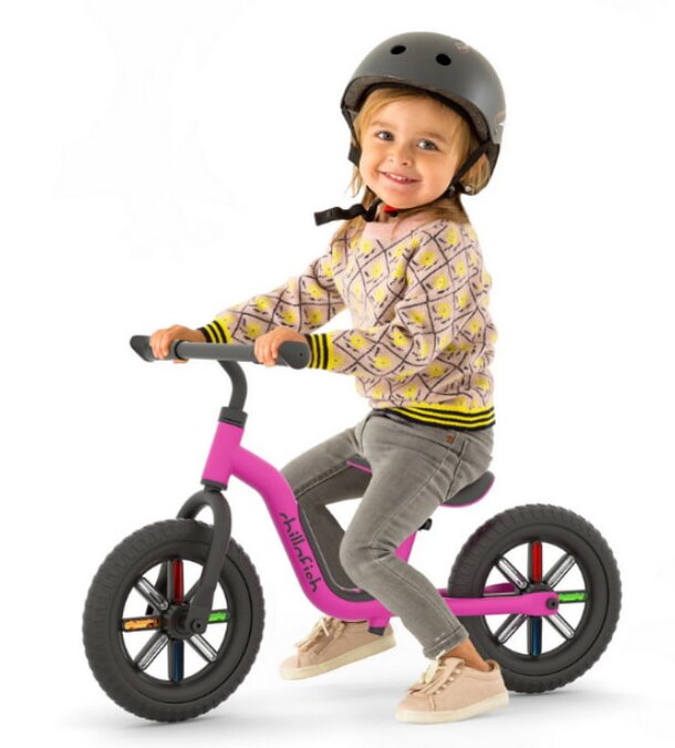 A little girl in a helmet rides a pink bike, smiling as she enjoys her outdoor adventure.