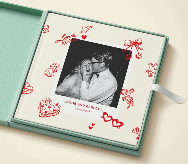 An open mint-green box contains a photo album decorated with red love-themed doodles. The cover photo shows a smiling couple, and text reads "Jacob and Rebecca, 14.02.2025."