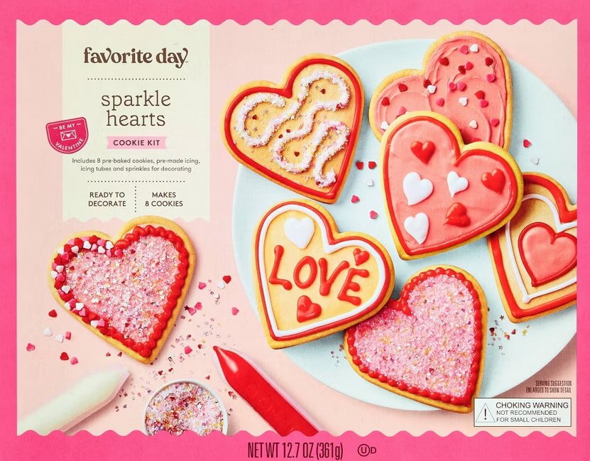 Heart-shaped cookies with pink, red, and white icing and sprinkles on a plate, alongside icing tubes. The tone is festive and playful.
