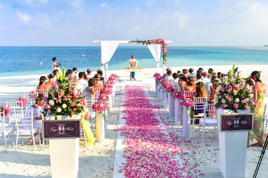 Beach wedding setup with rows of white chairs and pink floral arrangements, aisle covered in pink petals, and turquoise sea under a sunny sky.