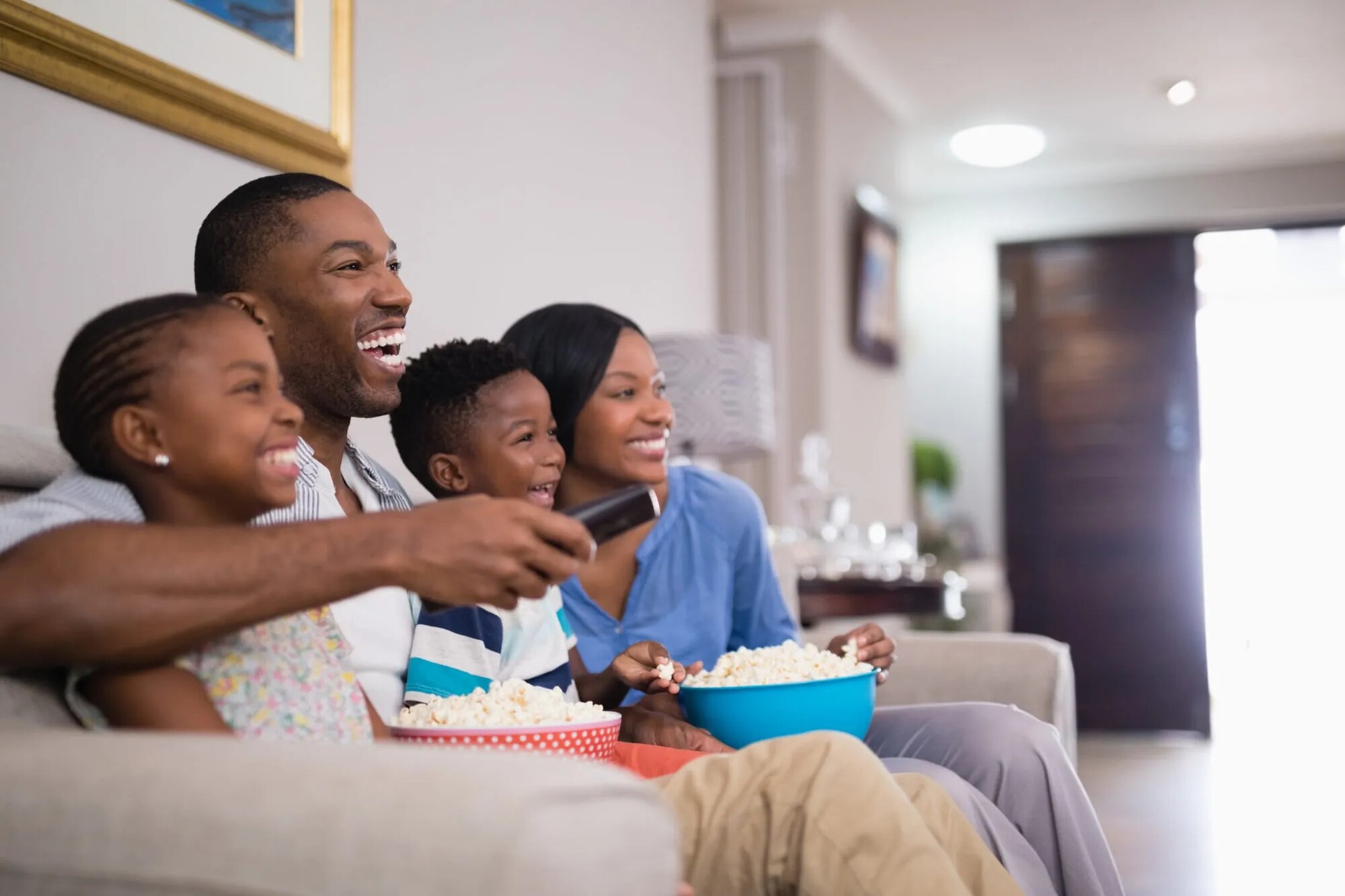 A family of four sitting together on a couch, watching television in a cozy living room setting.