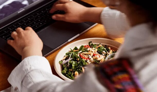 A woman types on a laptop while enjoying a salad, balancing work and a healthy meal simultaneously.