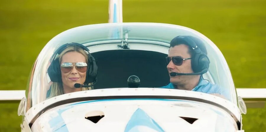 Two pilots wearing headsets and sunglasses sit in a small airplane cockpit, with a grassy field visible through the curved windshield, conveying a sense of adventure.