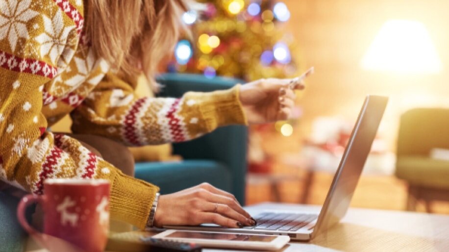 A woman in a sweater works on a laptop, seated in front of a beautifully decorated Christmas tree.