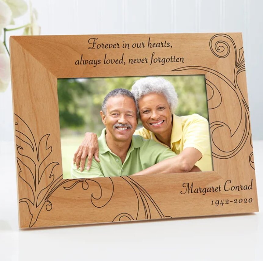 A wooden frame displays a smiling elderly couple in a field. Engraved text reads, “Forever in our hearts, always loved, never forgotten. Margaret Conrad 1942-2020.” The tone is warm and loving.