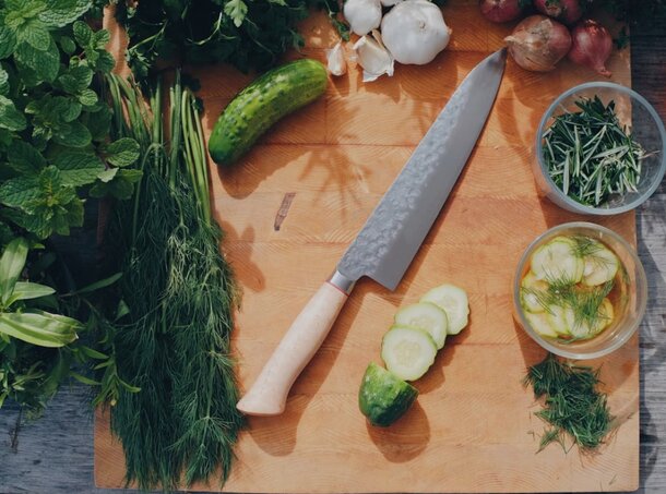 A wooden cutting board with a knife and sliced cucumbers alongside various chopped vegetables.
