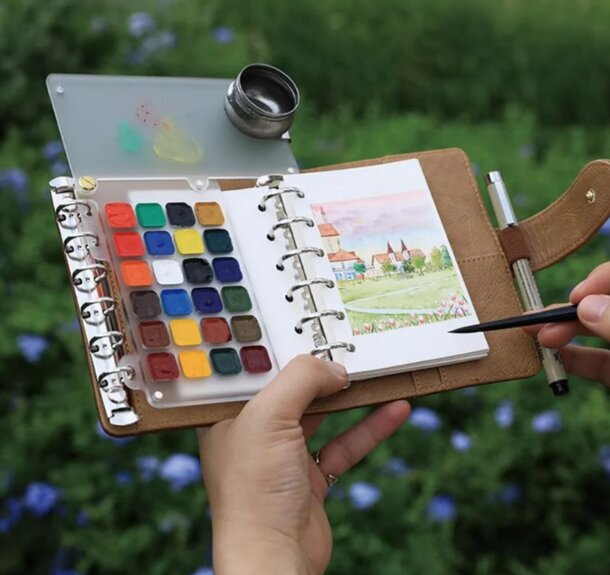 A person holds a watercolor notebook and a paintbrush, ready to create art outdoors.