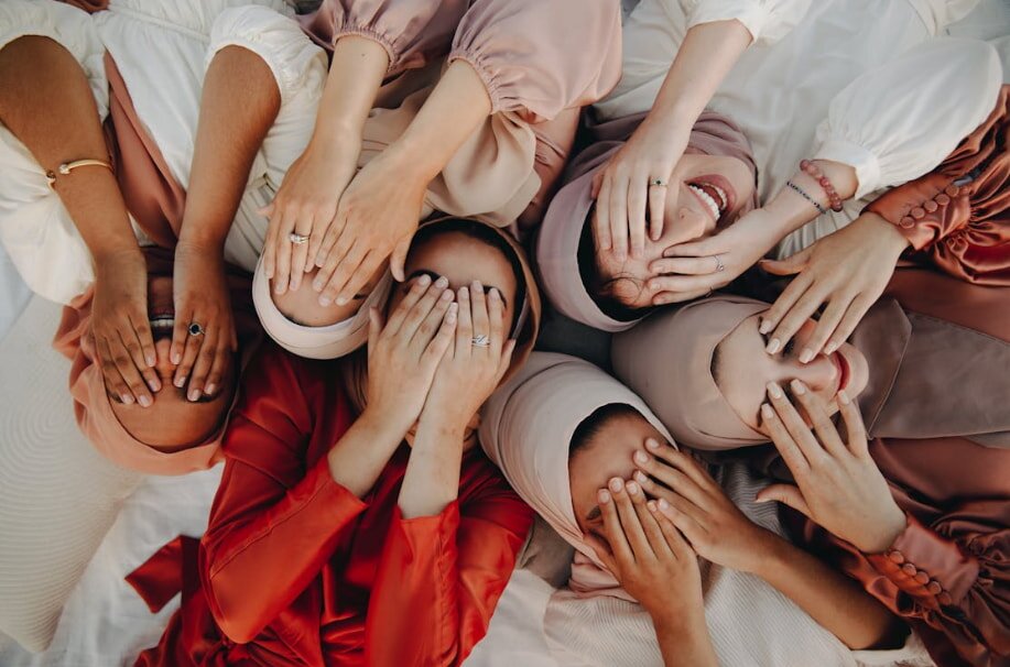 Six women in pastel and red outfits lie on a bed, covering their faces with their hands, conveying joy and unity in a playful manner.