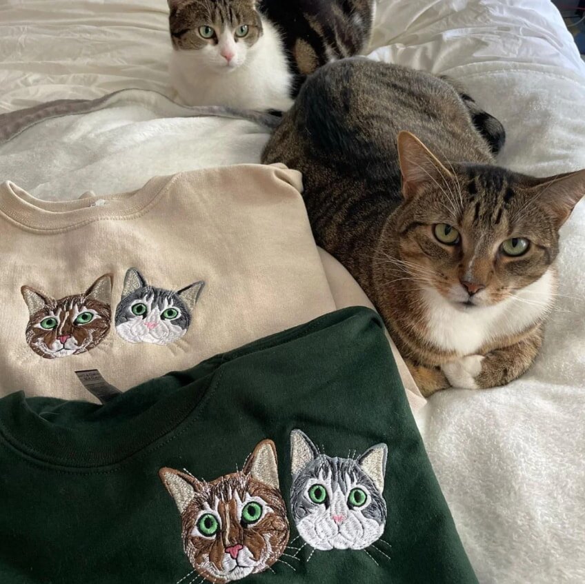 Two cats sit on a bed next to sweatshirts featuring embroidered cat faces resembling them. The setting is cozy, with a playful and warm tone.