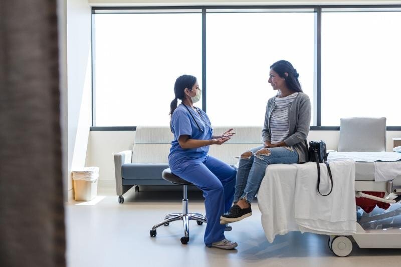 A woman in scrubs converses with another woman in a hospital room, discussing care or treatment options.