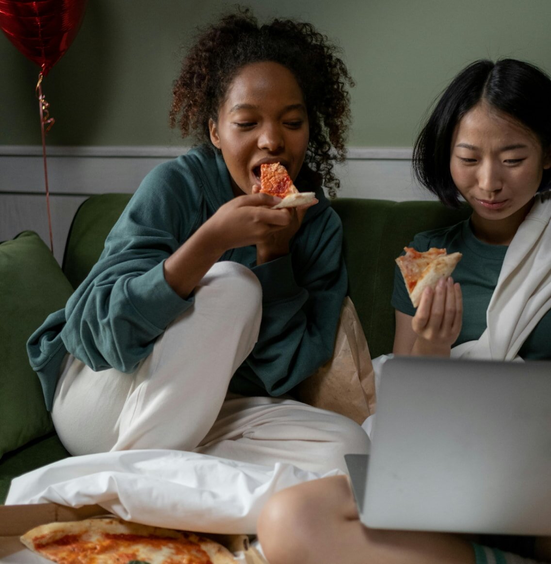 Two women share a pizza on a couch, smiling and enjoying their meal together.