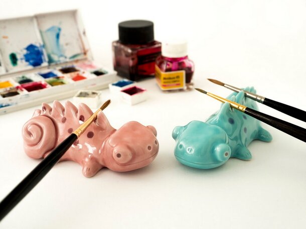 Two small ceramic animals sit beside a set of colorful paint brushes on a wooden surface.