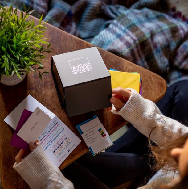 A woman sits at a wooden table, examining a box of cards placed in front of her.