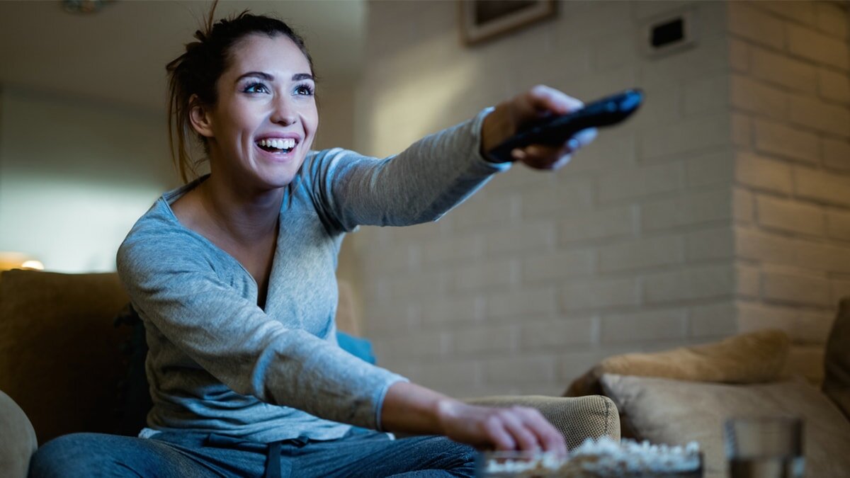 A woman sits on a couch, holding a remote control, appearing relaxed and engaged in her surroundings.
