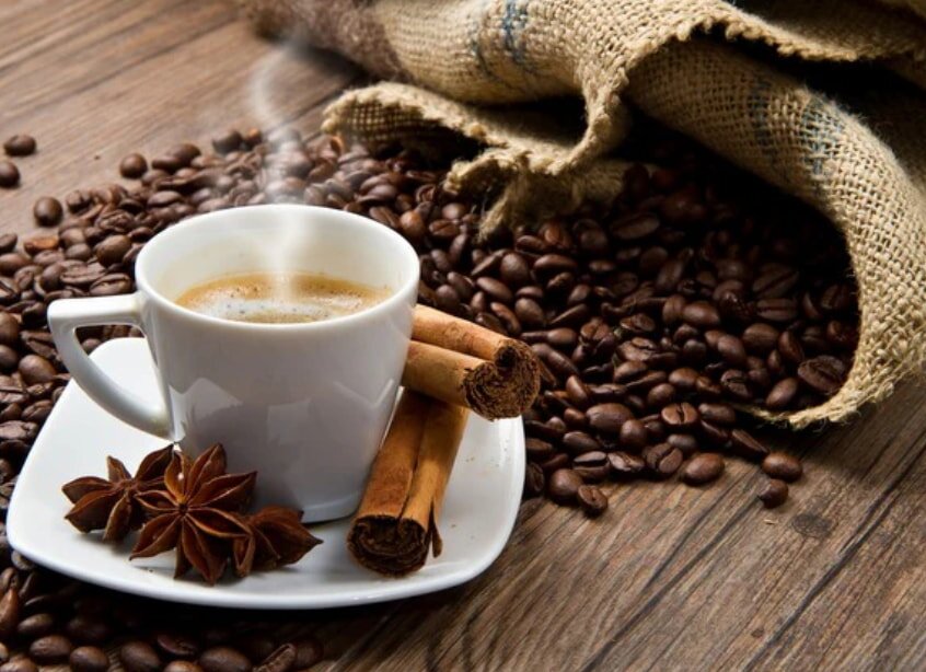 Steaming coffee in a white cup on a saucer, surrounded by coffee beans, cinnamon sticks, and star anise. A burlap sack lies open nearby.
