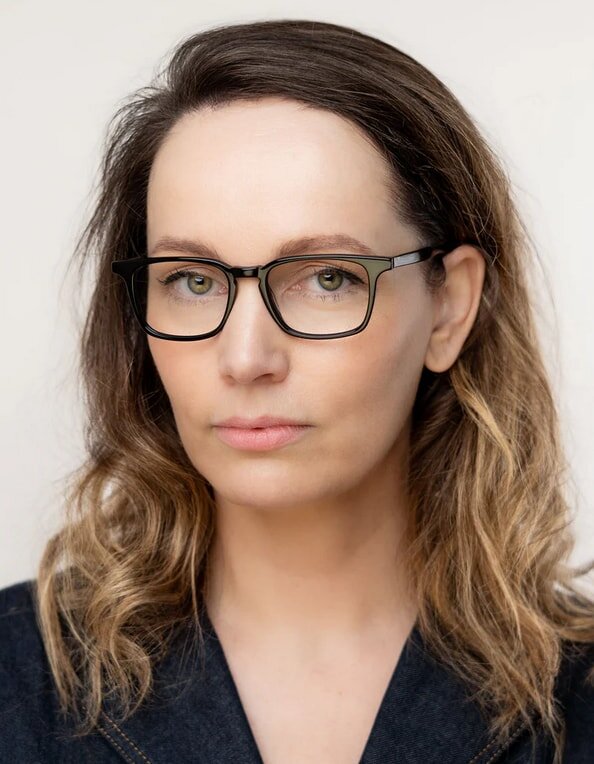 A woman with glasses and wavy hair looks confidently at the camera, wearing a dark jacket against a plain background.