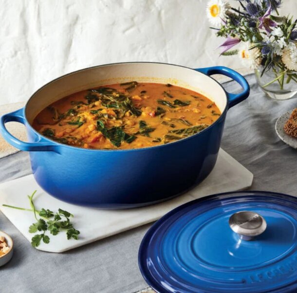 A blue Dutch oven beside a bowl of soup and a plate of colorful vegetables on a wooden table.