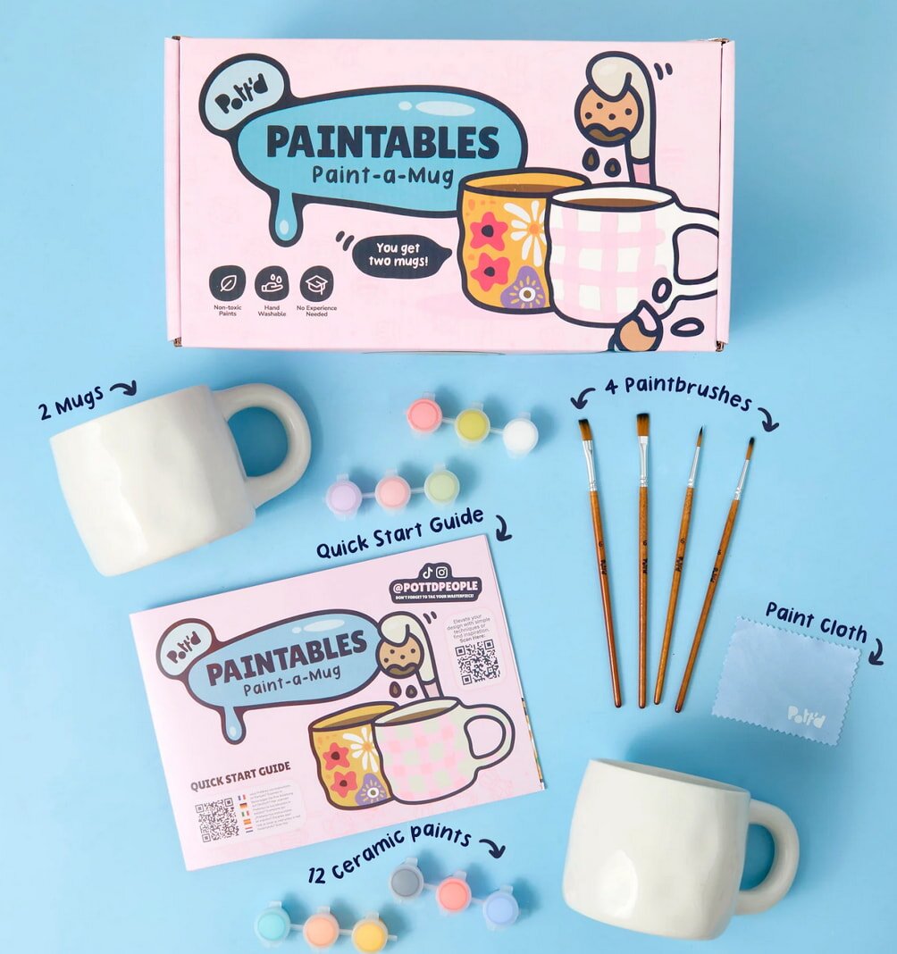 A "Paintables" mug kit displayed on a blue background includes two white mugs, 12 colorful ceramic paints, four paintbrushes, a quick start guide, and a paint cloth. The packaging is pink with whimsical illustrations, creating a fun and creative tone.